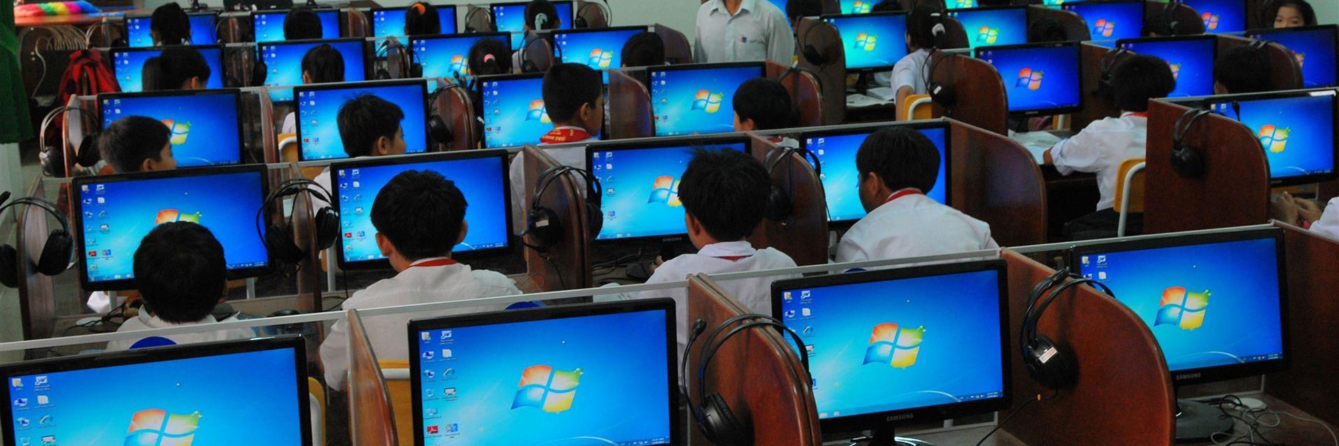Schüler lernen in einem Computerraum mit mehreren Monitoren und einem lehrenden Lehrer.